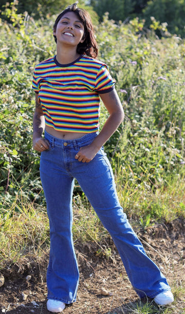 Meera is stood in a grass field wearing the stone wash blue stretch denim rock n roll bell bottom flares with a retro rainbow stripe short sleeved tshirt and white trainers. She is facing forward posing with one leg kicking forward and both hands resting on the front jean pockets whilst smising to camera.