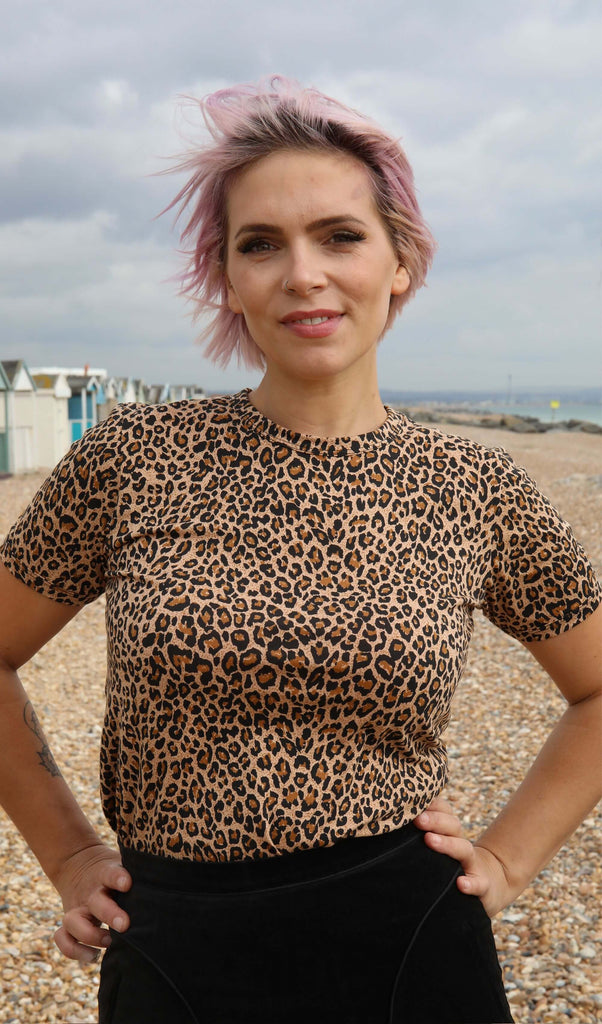 white femme model with short pink hair is stood on Brighton seafront wearing Natural All Over Leopard Print T Shirt posing toward the camera with her hands on her hips. 