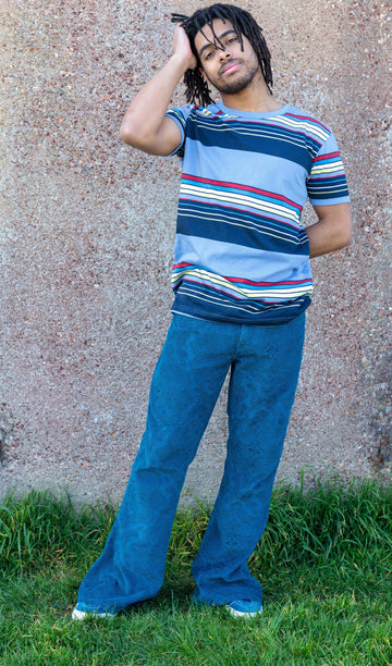 Basil is stood outside in front a brown wall wearing the ink blue paisley corduroy bell bottom super flares with a retro blue striped tshirt and blue trainers. He is posing facing the camera with one leg out to highlight the flare and is posing toward the camera with one hand in his hair.