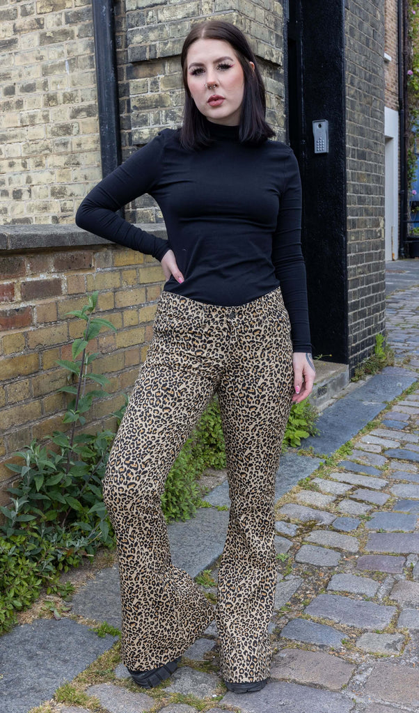 Model with dark hair wearing Leopard Print Bell Bottom Stretch Super Flares, paired with a long sleeve black turtleneck top and chunky black boots. The flares are a natural leopard print pattern and colour. Model is posing with one hand hip and the other by her side.