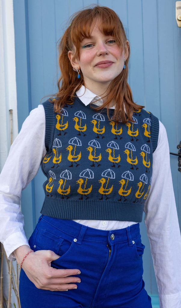 Model with red hair wearing Nice Weather for Ducks Cropped Knitted Tank Top paired with yellow hair accessories, blue earrings, white shirt and bright blue jeans. The tank top is a dusty dark blue colour with rows of yellow ducks holding white umbrellas printed on it. The tank top is a cropped style. Model is looking at camera and smiling