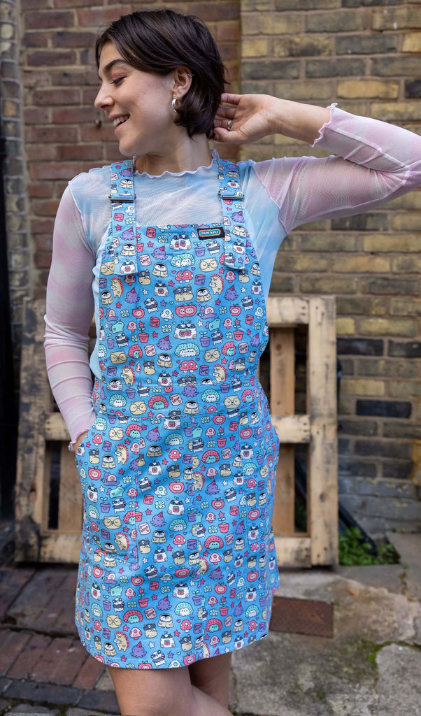 Freya, a white femme model with short brown hair, is stood outside in Hove wearing Run & Fly x Sugar & Sloth Stretch Twill Pinafore with a pastel mesh top underneath. The baby blue pinafore dress has an all over character print illustrated by Sugar & Sloth. Freya is smiling and facing the camera with one hand in the pinafore pocket and other in her hair. 
