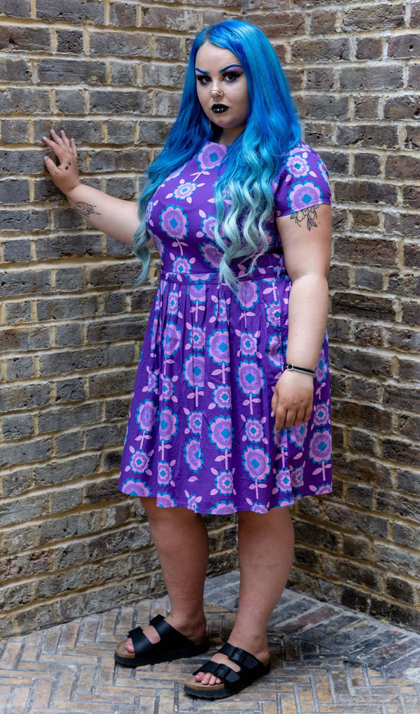 a white femme model with long blue hair and dark makeup is stood in front of a brick wall in Hove wearing Purple Retro Flowers Belted Stretch Tea Dress with Pockets with black sandals. The model is posing with one hand leant against the wall and looking at the camera. 
