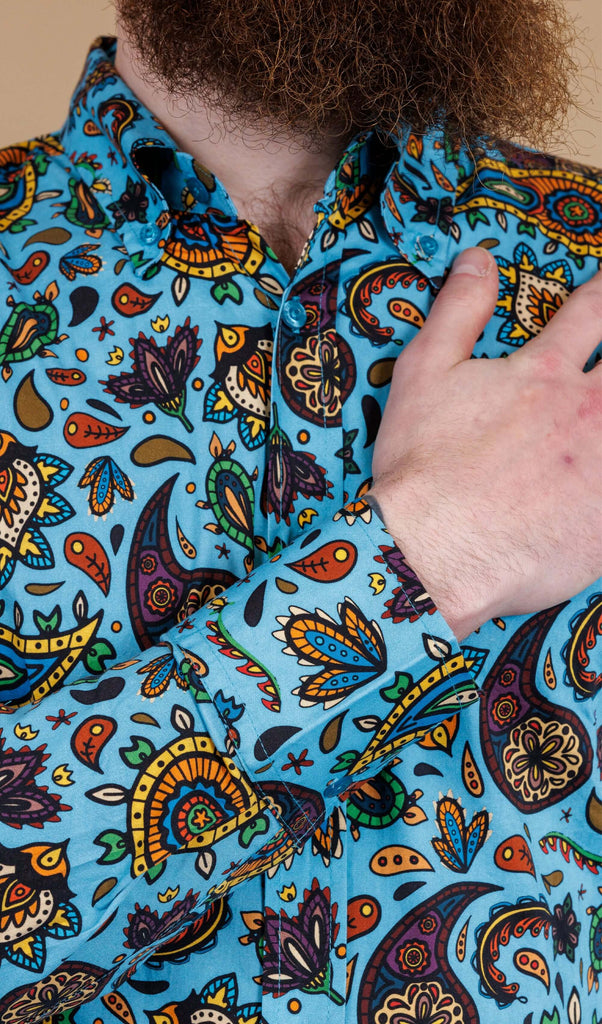 Close up of Dec wearing Sky Blue Paisley Print Long Sleeve Shirt. The shirt is a sky blue colour with an all over paisley print in various muted rainbow colours. Dec's hand is resting on his chest.