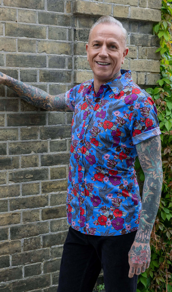 James, a tattooed male model, is stood outside in Hove wearing Hummingbird Print Short Sleeve Shirt with black trousers. The shirt is blue with an all over pattern of hummingbirds with red and purple flowers. James is smiling facing the camera with one hand leaning in a brick wall.