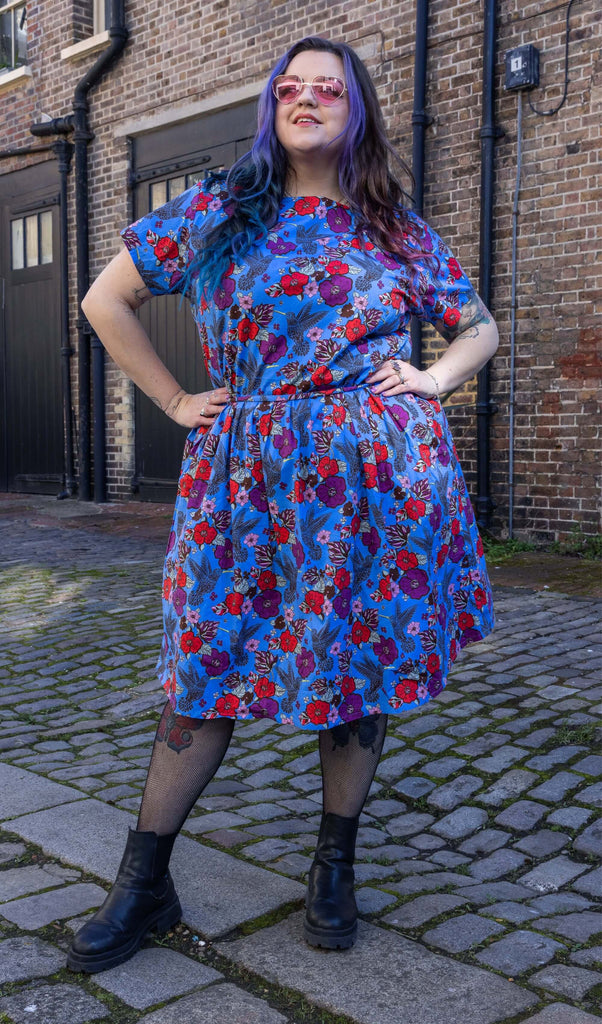 Luisa, a femme model with purple hair is stood outside in Hove wearing Hummingbird Stretch Belted Tea Dress with Pockets paired with fishnet tights, black boots and pink heart shaped sunglasses. The dress is blue with an over hummingbird print with red and purple flowers. Luisa is posing towards the camera with her hands on her hips in the sun. 