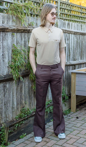 Jack is stood outside in a garden wearing the Presley brown bell bottom trouser slacks with a short sleeve sand coloured short sleeved shirt and white trainers. They have mid length blonde hair and round glasses. They are facing the camera posing with both hands in the trouser pockets and looking off to the right.