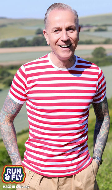 Jim is stood outside in a field wearing the white and red stripe short sleeve tshirt with beige cargos. They are facing forward posing with both hands in their front trouser pockets whilst smiling to camera. Photo is cropped from the hips up.