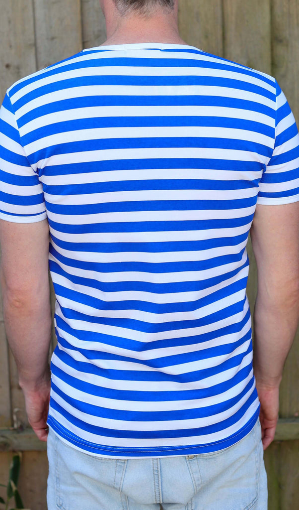 male model is stood outside in front of a wooden fence wearing the Royal & White Stripe Short Sleeve T Shirt with light blue jeans. He is stood with his back to the camera and arms resting by his sides.
