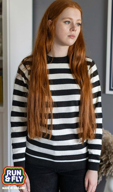 Sophie is stood in front of a grey background in front of a plant and has long red hair. They are wearing the White and Black Striped Jumper with dark jeans.
