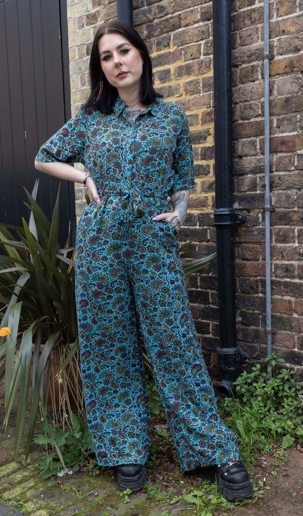 Tattooed model with dark hair wearing Sky Blue Paisley Jumpsuit, paired with chunky black boots. The jumpsuit is a sky blue colour with an all over paisley print in various muted rainbow colours. There is a matching fabric belt which is tied around model's waist in a bow at the front. Model is posing with one hand on hip and other hand in jumpsuit pocket.