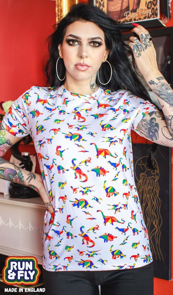  Rosie is stood in front of a red wall containing framed prints wearing the white rainbow dinosaur tshirt with black trousers. They are facing forward posing with one hand on their hip whilst the other running through their hair. Photo is cropped from the hips up.