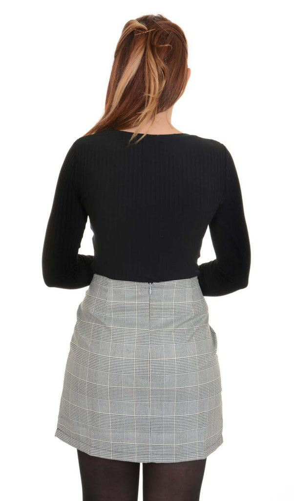 Model is stood in front of a white studio background wearing the retro black and white prince of Wales mini skirt with a long sleeve black tshirt and tights. They are facing away from the camera with their arms crossed in front of them. Photo is cropped from the knees up.
