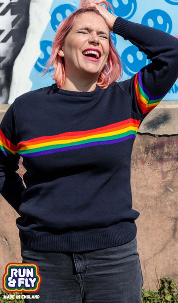 Alex is stood leaning against a graffiti wall wearing the rainbow jumper with dark denim jeans. She is facing forward posing with one hand pushing back her hair and the other arm behind her whilst laughing. Photo is cropped from the hips up.
