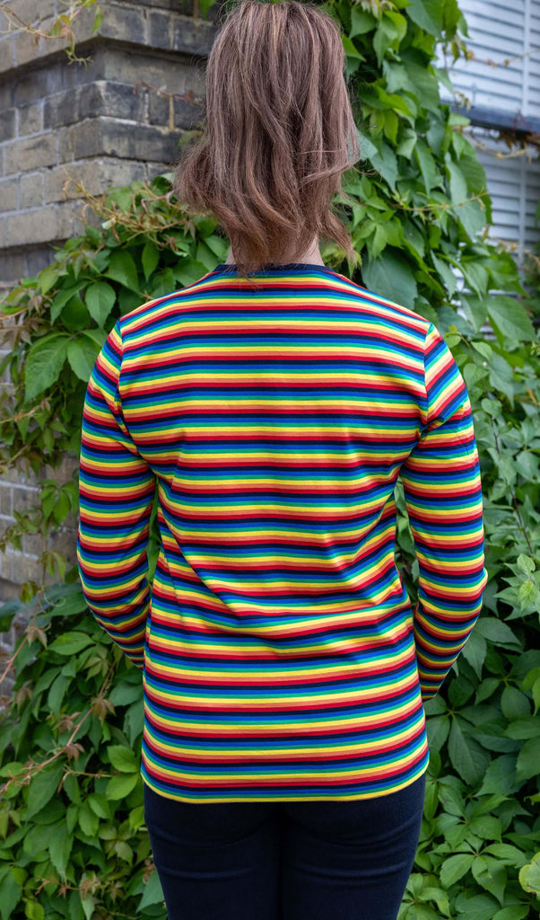 Sophie is wearing Retro Rainbow Brights Repeat Striped Long Sleeved T Shirt with black trousers. She is stood outside in Hove in front of a brick wall with greenery with her back to the camera.