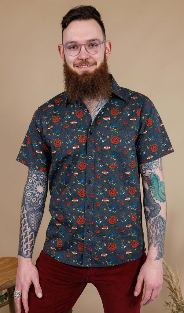 Dec, a white male model with a beard and glasses is wearing Retro Old School Tattoo Print Short Sleeve Shirt with Tawney Port Corduroy Bell Bottom Flares. The print has traditional tattoo style red roses, blue swallows, yellow envelopes, anchors and valentines love heart. Dec is stood in front of a beige backdrop at a photography studio in Hove facing the camera and smiling with his hands by his sides.