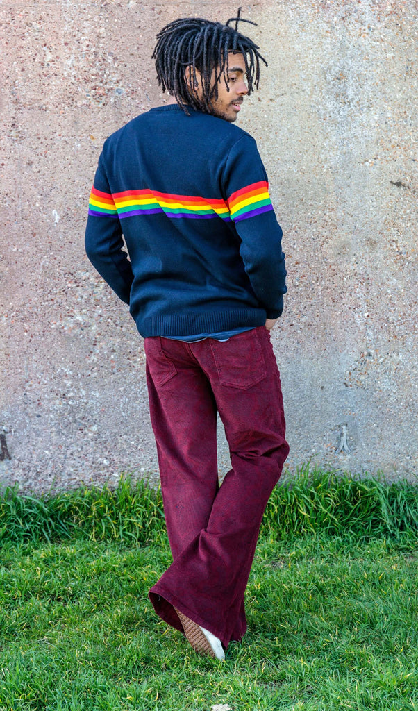 Basil is stood in front of a plain brick wall wearing the tawney port paisley corduroy bell bottom super flares with the navy retro rainbow stripe jumper and blue trainers. He is facing away from the camera taking a step forward and looking over his shoulder.
