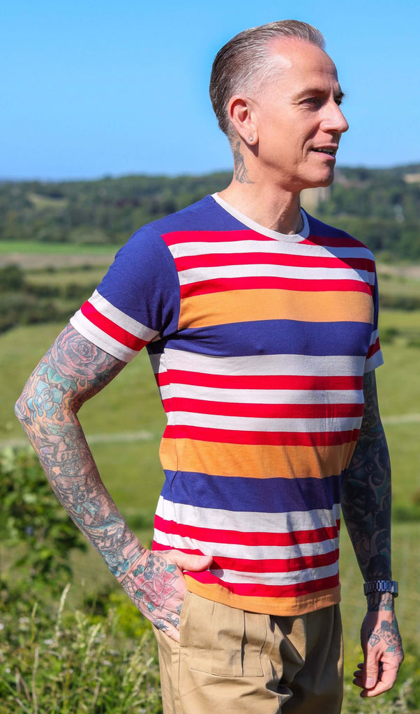 Jim is stood on a grass hill wearing the retro multi striped nugget tshit with beige cargo trousers. They are heavily tattooed with swept back grey hair. They are facing to the right with one hand in their trouser pocket smiling to the right. Photo is cropped from the hips up.