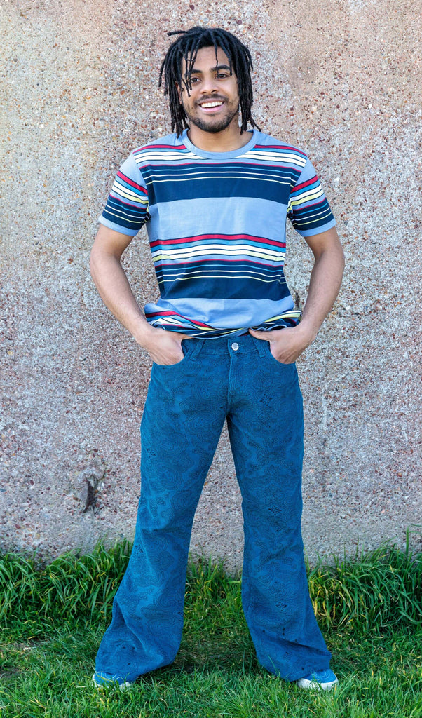 Basil is stood outside in front a brown wall wearing the ink blue paisley corduroy bell bottom super flares with a retro blue striped tshirt and blue trainers. He is posing facing the camera with one leg out to highlight the flare and both hands in the front pockets, he is smiling towards the camera.