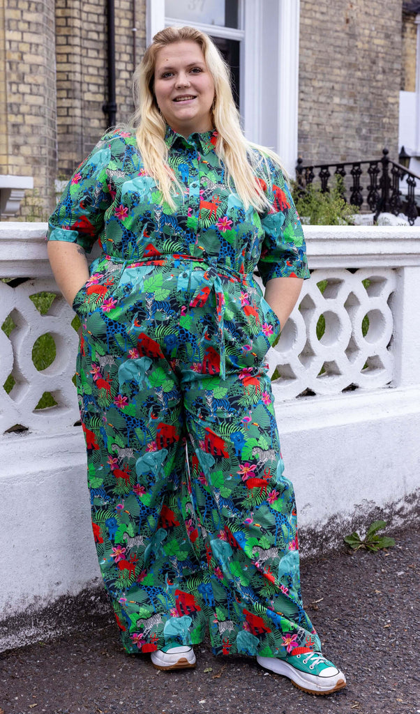 Model with long blonde hair wearing Jungle Stretch Jumpsuit, paired with teal coloured trainers. The jumpsuit is a forest green colour with all over print of various jungle animals and plants in black, white, red, pink, yellow and blue. The collared jumpsuit is buttoned up to the top with matching fabric belt tied around the waist. The model is stood outside in Hove smiling towards the camera with her hands in the jumpsuit pockets.