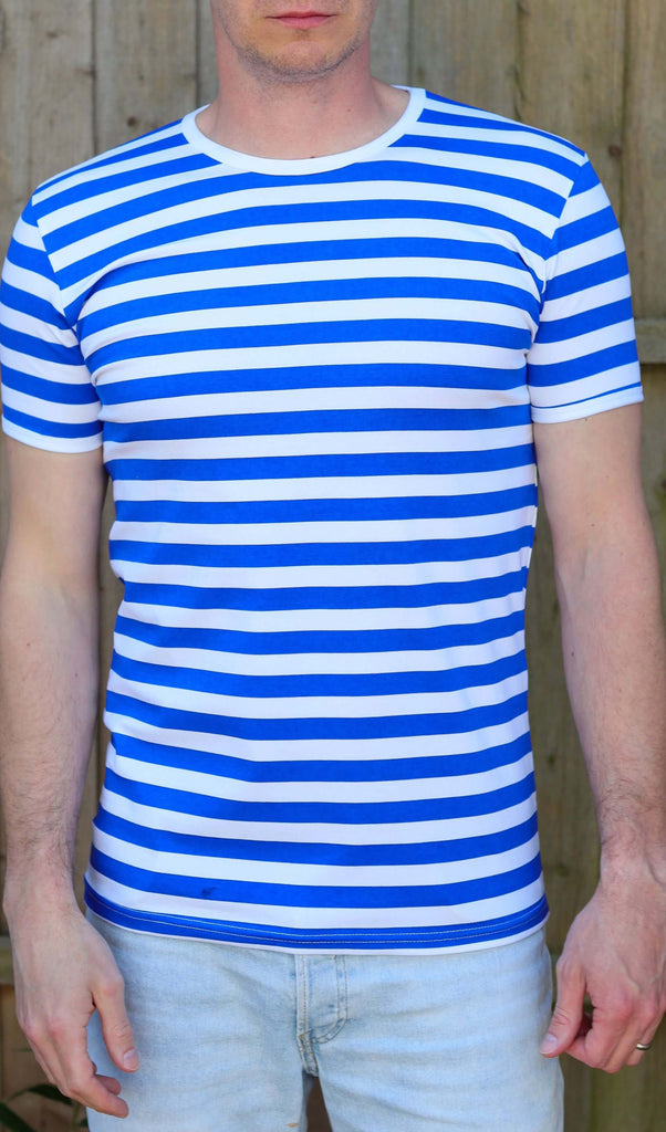 male model is stood outside in front of a wooden fence wearing the Royal & White Stripe Short Sleeve T Shirt with light blue jeans. He is facing the camera with his arms resting by his sides.