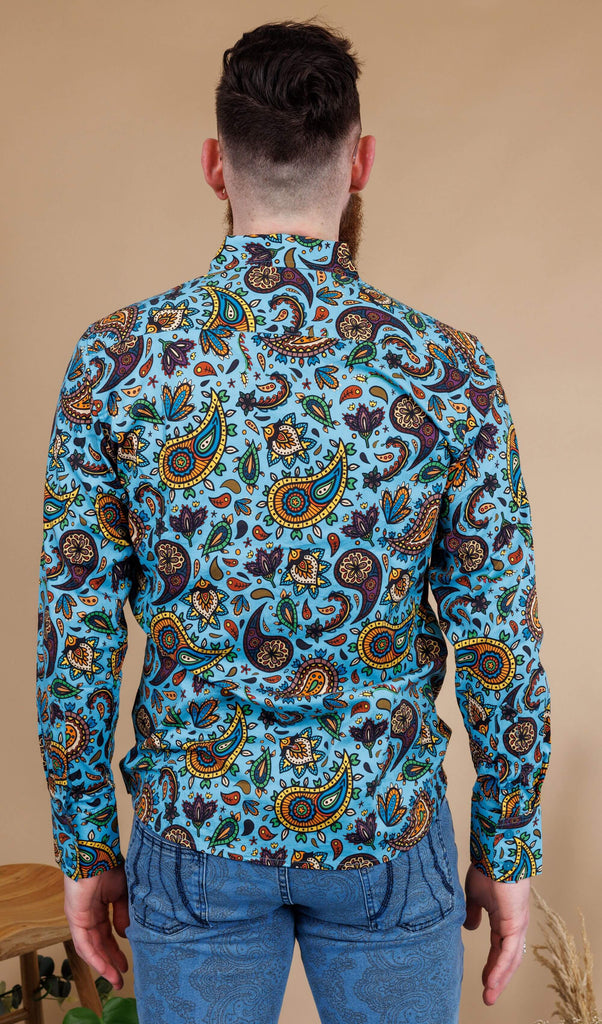 Dec is stood in a photography studio in Hove in front of a beige backdrop wearing Sky Blue Paisley Print Long Sleeve Shirt with blue jeans. The shirt is a sky blue colour with an all over paisley print in various muted rainbow colours. He is stood with his back to the camera and his arms down by his sides. 