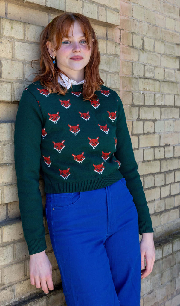  Model is wearing Green Fox Cropped Jumper, paired with a white collared shirt and bright blue jeans. The jumper is a dark forest green colour with a print of multiple red foxes on the front torso. Model is leant against a wall, smiling at camera.