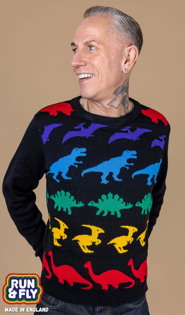 James is wearing Rainbow Dinosaur Mash Up Jumper with jeans. The jumper is a plain black base with various dinosaurs in a rainbow colour stripe sequence with plain black arms and back. James is stood in front of a beige backdrop posing with his hands behind him and smiling looking off to one side. 
