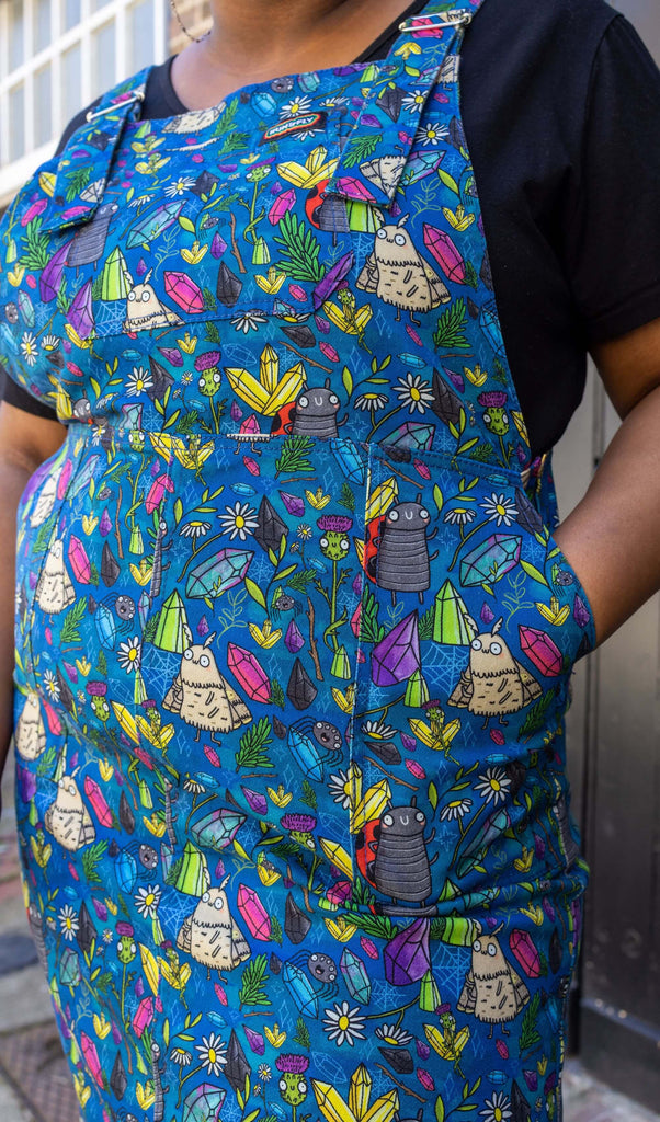 close up of Han's hand in the pocket of the Run & Fly x Katie Abey Crystal Critters Stretch Twill Long Pinafore Dress. The blue pinafore dress has an all over print of critters and crystals illustrated by Katie Abey.
