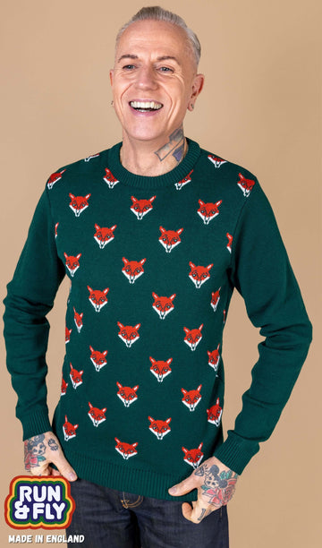 a white male tattooed model is stood in front of a beige backdrop wearing Forest Green Fox Head Jumper paired with jeans. The green long sleeved jumper has an all over print of fox heads on the torso. The model is smiling whilst holding the bottom of the jumper. 