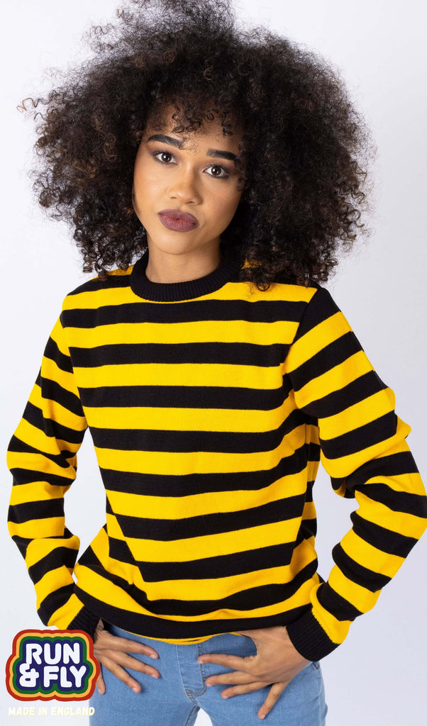 Cindy is stood in front of a white studio background wearing the yellow and black striped jumper with blue denim jeans. They are facing forward posing with both hands resting on their front jean pockets. Photo is cropped from the hips up.