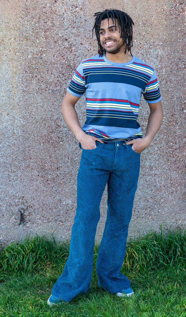 Basil is stood outside in front a brown wall wearing the ink blue paisley corduroy bell bottom super flares with a retro blue striped tshirt and blue trainers. He is posing facing the camera with one leg out to highlight the flare and both hands in the front pockets, he is looking off to the left and smiling.