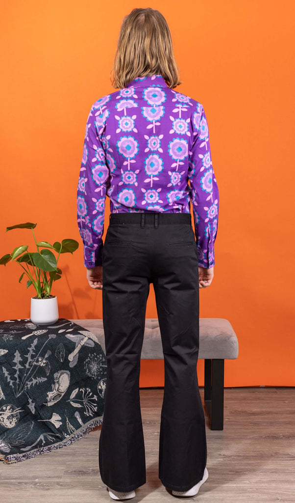 Jack is wearing Presley Black Stretch Cotton Twill Bell Bottom Trouser Slacks paired with Purple Retro Flowers Long Sleeve Shirt and white trainers. Jack is posing in a photography studio in Hove with an orange backdrop and his back to the camera.