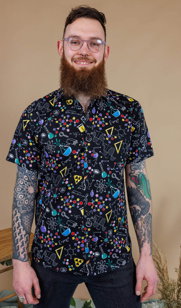 Dec is wearing the science print short sleeve shirt, featuring multicoloured science symbols on black. Model is posing in studio with beige brown backdrop and smiling, at the camera with hands by sides.