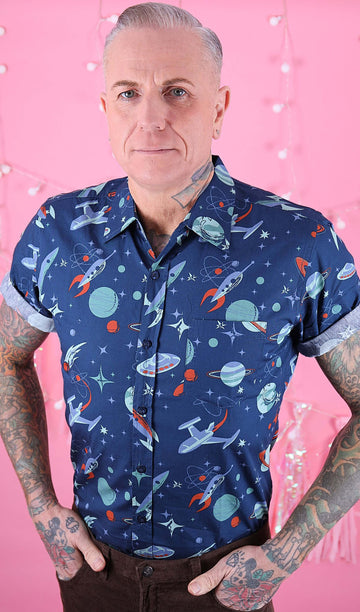 Jim is stood in front of a pink studio background wearing the outer space retro short sleeve shirt with brown corduroy trousers. He is heavily tattooed with silver swept back hair. He is posing facing the camera with both hands in his front trouser pockets. Photo is cropped from the hips up.