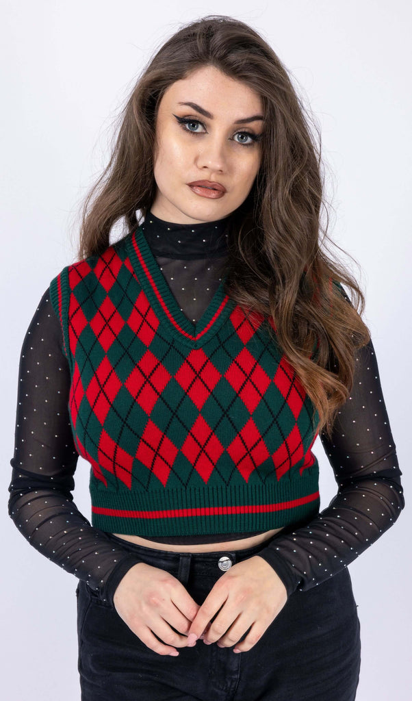 Charlotte is wearing Red and Green Argyle Knitted Tank Top with a long sleeve black mesh top underneath with white polka dots and black trousers. Charlotte is stood in front of a white background posing toward the camera with her hands together. The photo is cropped from the thighs down. 