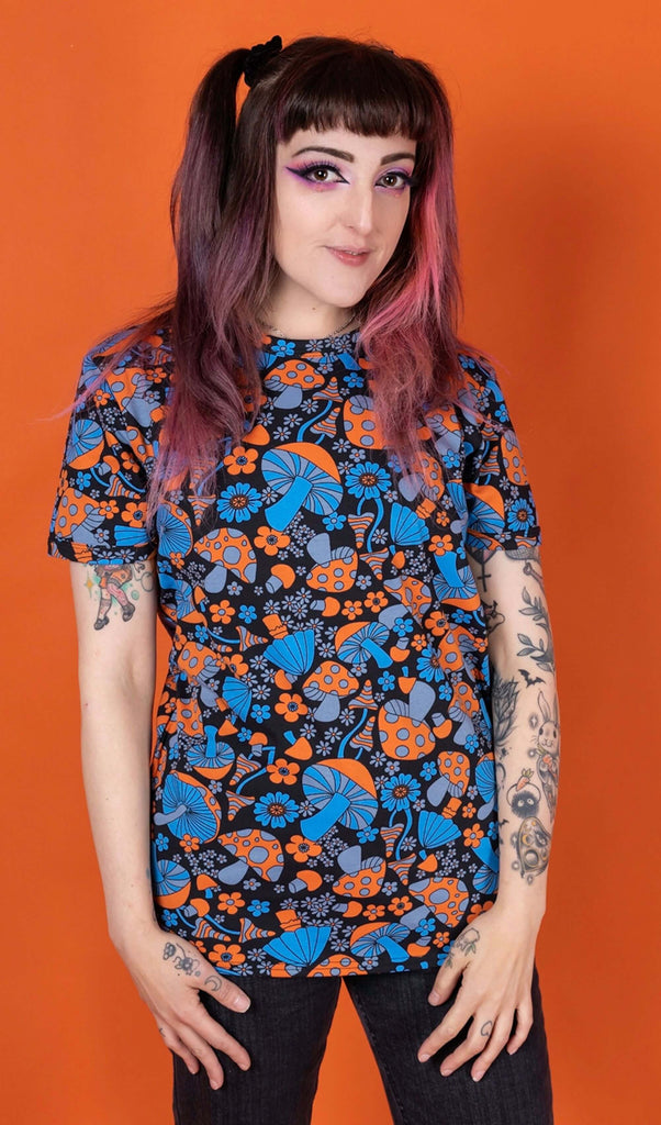 Sarah is stood in front of an orange background wearing the Magic Mushroom Short Sleeve Tee with black stonewash denim flares. She is facing forward with both hands resting on her hips whilst smiling to camera. The magic mushroom print is a black base tshirt with blue and neon orange toadstool mushrooms and daisy shape flowers all over.