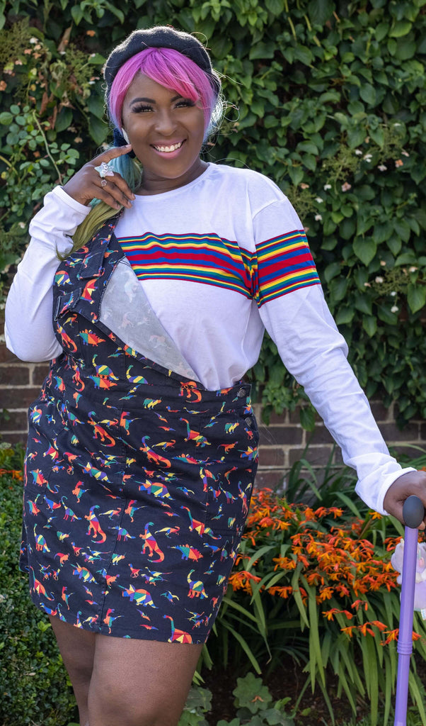 Ayesha smiling, holding a bright purple walking stick and wearing the Retro Rainbow Engineered Striped Long Sleeve T Shirt with a rainbow dino pinafore dress