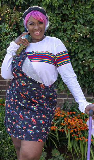 Ayesha smiling, holding a bright purple walking stick and wearing the Retro Rainbow Engineered Striped Long Sleeve T Shirt with a rainbow dino pinafore dress