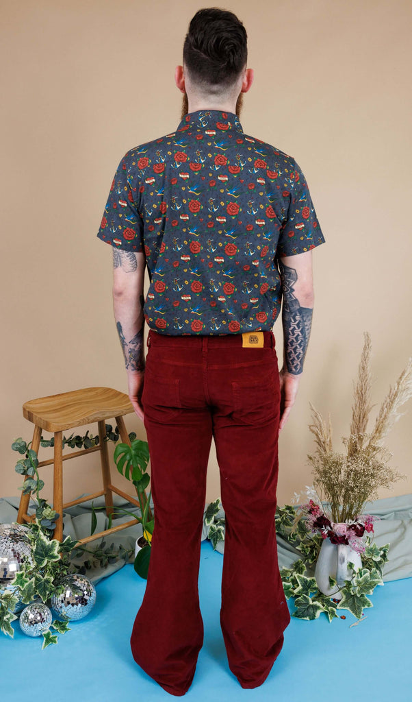 Dec, a white male with a beard and glasses, is stood in a photography studio in Hove in front of a beige backdrop wearing Tawney Port Corduroy Bell Bottom Flares with Retro Old School Tattoo Print Short Sleeve Shirt. Dec is stood with his back to the camera and arms down by his sides.