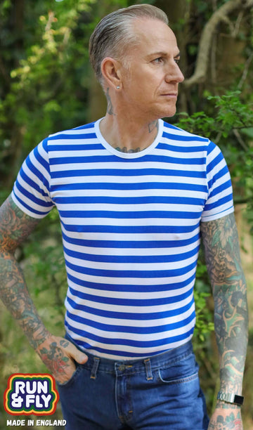 tattooed male model is stood outside amongst trees wearing the Royal & White Stripe Short Sleeve T Shirt with blue jeans. He is posing facing the camera and looking off to one side with one hand in his pocket and other resting by his side.