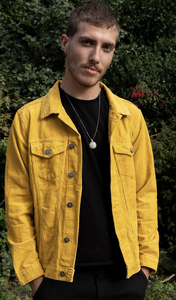 Jake is stood in a garden area wearing the retro vintage gold cord unisex western jacket with a black tee and black trousers with a pendant necklace. He is facing the camera smiling with both hands in his front jean pockets. Photo is cropped from the hips up.