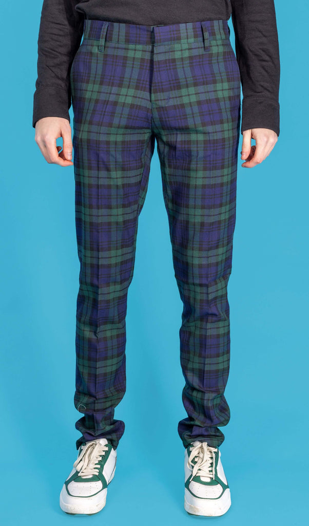 Jack is stood in front of a blue studio background wearing the retro mod stretch black watch tartan trousers with a long sleeve black tshirt and white trainers. They are facing forward posing with both arms resting by their side. Photo is cropped from the waist down.