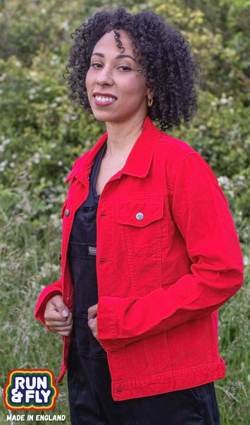 Amy is stood outside in a field area wearing the retro vintage red cord unisex western jacket with black dungarees. They are facing to the left looking over to the camera smiling whilst pulling the jacket together. Photo is cropped from the hips up.