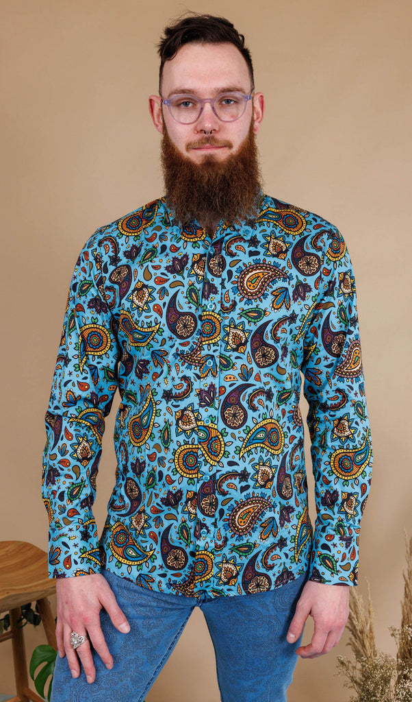 Dec is stood in a photography studio in Hove in front of a beige backdrop  wearing Sky Blue Paisley Print Long Sleeve Shirt with blue jeans. The shirt is a sky blue colour with an all over paisley print in various muted rainbow colours. He is facing the camera and smiling with his arms by his sides.