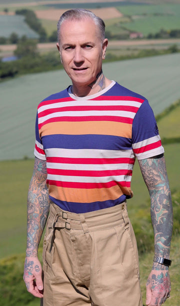 Jim is stood on a grass hill wearing the retro multi striped nugget tshit with beige cargo trousers. They are heavily tattooed with swept back grey hair. They are facing forward posing with both arms out by their sides whilst smiling to camera. Photo is cropped from the hips up.