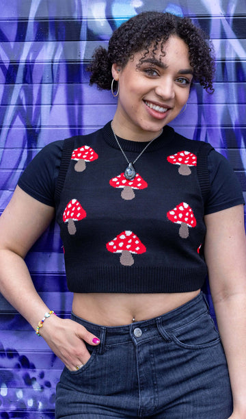  Georgia is stood in front of a graffiti wall wearing the mushroom knitted tank top over a black short sleeve shirt and black jeans. She's posing facing the camera smiling with one hand in the front jean pocket. Photo is cropped from the hips up.