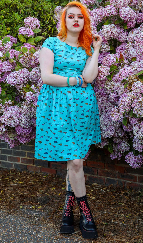 Tasha is stood in front of a floral bush wearing the tartan dinosaur tea party dress with black and red boots. She has mid length bright orange hair and various tattoos. She is facing forward posing with her legs crossed and one hand resting across her middle and the other resting by her face.