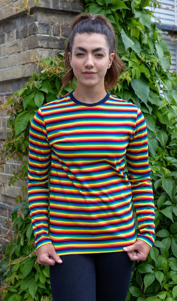 Sophie is wearing Retro Rainbow Brights Repeat Striped Long Sleeved T Shirt with black trousers. She is stood outside in Hove in front of a brick wall with greenery and is posing facing the camera while holding the bottom of the t shirt.