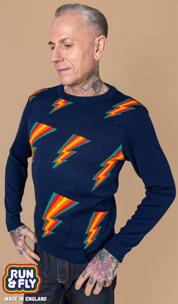 James is stood in front of a beige background wearing the run and fly navy lightning bolt jumper with denim jeans. He is facing left with both hands on his hips whilst looking down. The jumper is a navy blue base colour with repeating green, red, orange and yellow lightning bolts on the chest. The arms and back are plain navy blue. The photo is cropped from the hips up. In the left corner is a Made in England logo.